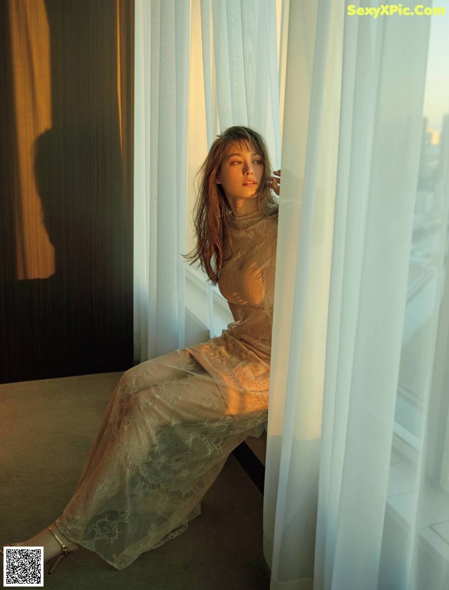 A woman in a long dress sitting by a window.